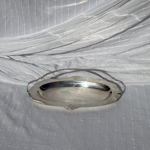 Op de afbeelding: Een ovaal zilverkleurig serveerblad met een gepolijste metalen afwerking en een decoratief boogvormig handvat. Het blad heeft een gladde, reflecterende kom met gedetailleerde filigraanversieringen aan de basis van het handvat.