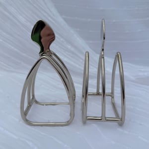May include: Two silver-colored metal napkin holders. One has a curved top with a decorative element, and the other has a simple, open design. The holders are made of thin, shiny metal and are set against a white background.