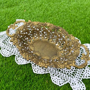 May include: An antique brass oval decorative basket with floral and scrollwork details. The basket has two handles and sits on a white lace doily. The basket is displayed on a green grassy surface.