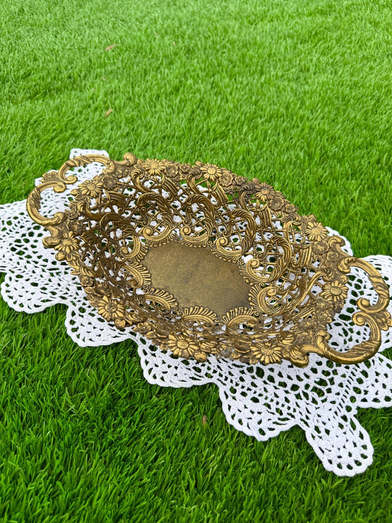 May include: An antique brass oval decorative basket with floral and scrollwork details. The basket has two handles and sits on a white lace doily. The basket is displayed on a green grassy surface.
