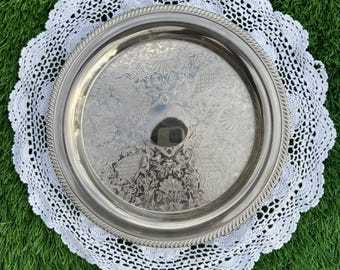 May include: A silver-colored, round serving tray with ornate embossed detailing, resting on a white, crocheted doily with a scalloped edge. The tray has a rope-like border. The background is green.