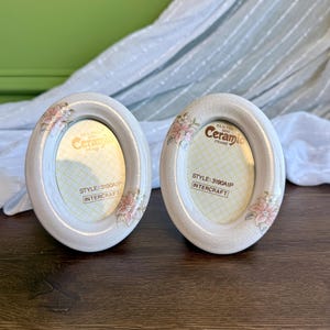 Puede incluir: Dos marcos de fotos ovalados de cerámica con un acabado de esmalte craquelado de color crema. Cada marco presenta detalles florales pintados en rosa y blanco. Los insertos muestran el texto: Kiln Fired Mini Ceramic Frame, Style: 3190A1P, Intercraft.