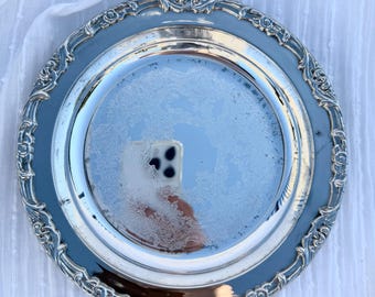 May include: A round, silver-colored serving tray with an ornate, floral-patterned border. The tray's center has a reflective surface with a subtle, etched design. The tray is likely made of metal and is suitable for serving food or displaying decorative items.