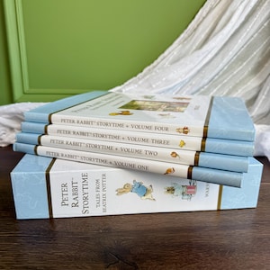 May include: A stack of four Peter Rabbit Storytime books resting on a matching box set. The spines are white with light blue accents, featuring the text Peter Rabbit Storytime Volume One through Four, Tales from Beatrix Potter, and Warn.