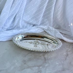 May include: An oval silver-toned serving tray featuring a polished, reflective surface with a decorative embossed floral motif in the centre and a scalloped, ridged edge detail along the entire perimeter of the dish.