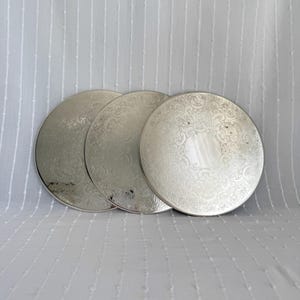 May include: Three silver-colored, round decorative plates with ornate, embossed floral patterns. The plates are arranged in a row, each displaying a detailed design. The background is a white, textured fabric.