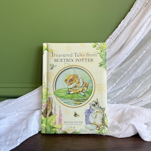 May include: A hardback book titled Treasured Tales from Beatrix Potter with a cream-coloured cover featuring a central circular illustration of a frog fishing on a lily pad, surrounded by botanical motifs and illustrations of small animals.