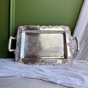 Puede incluir: Una bandeja de servicio rectangular de color plateado con detalles ornamentados en relieve de conchas y volutas a lo largo de los bordes. La bandeja incluye dos asas laterales integradas y una superficie central decorada con intrincados motivos florales grabados.