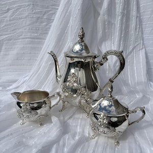 May include: A silver tea set comprising a teapot, creamer jug, and sugar bowl, is presented on a white backdrop. The set showcases intricate detailing and a reflective finish. The teapot has a decorative finial and a curved handle. The creamer and sugar bowl have small feet.