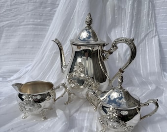 May include: A silver tea set, including a teapot, creamer, and sugar bowl, is displayed on a white surface. The set features ornate detailing and a reflective surface. The teapot has a decorative finial and curved handle. The creamer and sugar bowl have small feet.
