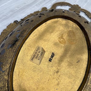 May include: A vintage, oval-shaped brass tray with ornate filigree edges and a textured surface. The tray has a gold finish and a label that reads "Made in Italy". The tray has a handle.
