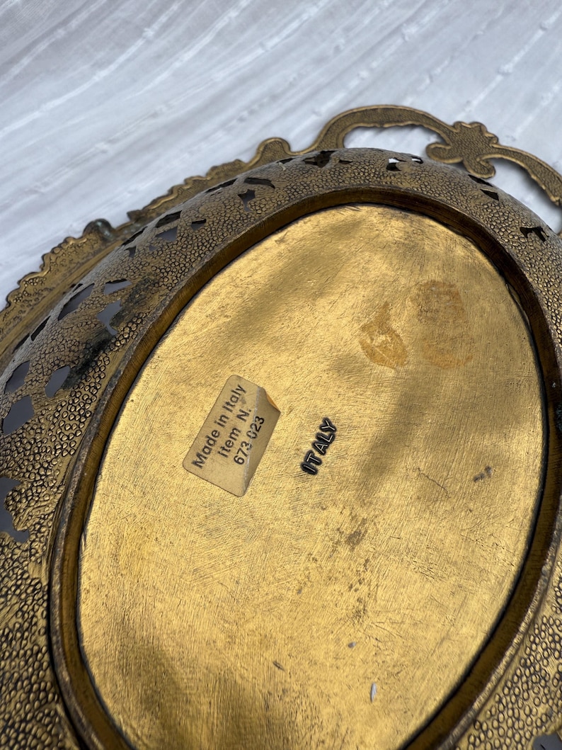 May include: A vintage, oval-shaped brass tray with ornate filigree edges and a textured surface. The tray has a gold finish and a label that reads "Made in Italy". The tray has a handle.