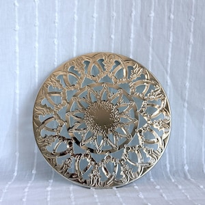 May include: A gold-coloured, circular decorative object with an intricate, openwork design. The design features a central sunburst pattern surrounded by ornate, swirling details. The object is set against a light blue background.