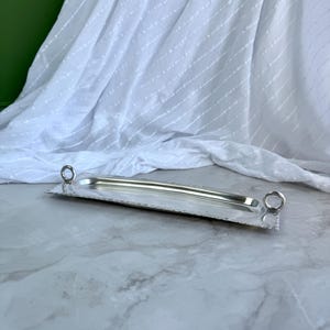 May include: A rectangular silver-toned metal serving tray featuring a smooth, recessed centre and a textured border. The tray includes two circular ring-shaped handles attached to each of the shorter ends, providing a functional grip for carrying.