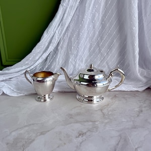 May include: A polished silver-toned teapot with a curved handle and a matching creamer jug. The teapot features a rounded body, a short spout, and a lid with a small knob. The creamer has a gold-toned interior and a pedestal base.