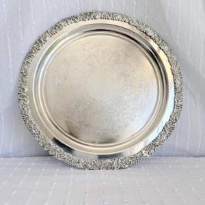 May include: A circular, silver-plated serving tray with an elaborate floral border. The tray's surface has a delicate, engraved floral design. The rim features a detailed, raised pattern. Suitable for serving food or displaying items.
