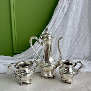 May include: A silver-plated coffee service set consisting of a tall coffee pot with a hinged lid and chain, a creamer, and a sugar bowl. Each piece features ornate embossed floral detailing, curved handles, and decorative feet on a polished surface.