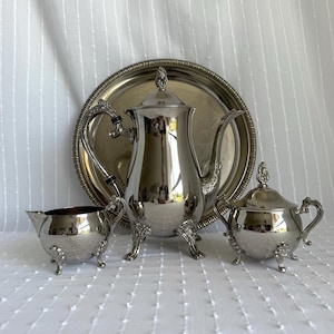 May include: A silver tea set comprising a teapot, creamer, sugar bowl, and a serving tray. The set features intricate detailing and is presented on a white surface. The teapot has a tall, curved spout and a decorative finial on the lid.