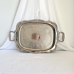 Può includere: Vassoio rettangolare argentato con manici decorati e un motivo floreale dettagliato. Il vassoio ha un bordo leggermente rialzato e un monogramma centrale. Un pezzo decorativo per servire.