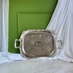 Puede incluir: Una bandeja de servicio rectangular de metal plateado con patrones florales y de volutas en relieve en el borde y las asas. La superficie central muestra un acabado deslustrado con intrincados grabados decorativos en el metal.