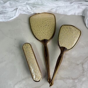 May include: A vanity set consisting of two hand mirrors and one hairbrush. Each piece features a metallic gold handle and a cream-coloured back panel with a textured, gold-toned lace-like pattern. The items have a matching design and finish.
