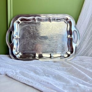 May include: A rectangular silver-toned serving tray featuring a scalloped edge, integrated side handles, and an intricate embossed floral pattern across the central surface. The metallic finish reflects light, highlighting the detailed engravings.