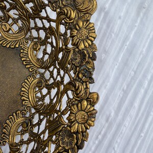 May include: Ornate gold-colored decorative plate with intricate floral and lattice detailing. The plate features a textured center and a detailed, raised border with flowers and swirling patterns. The design is symmetrical and the plate has an antique appearance.