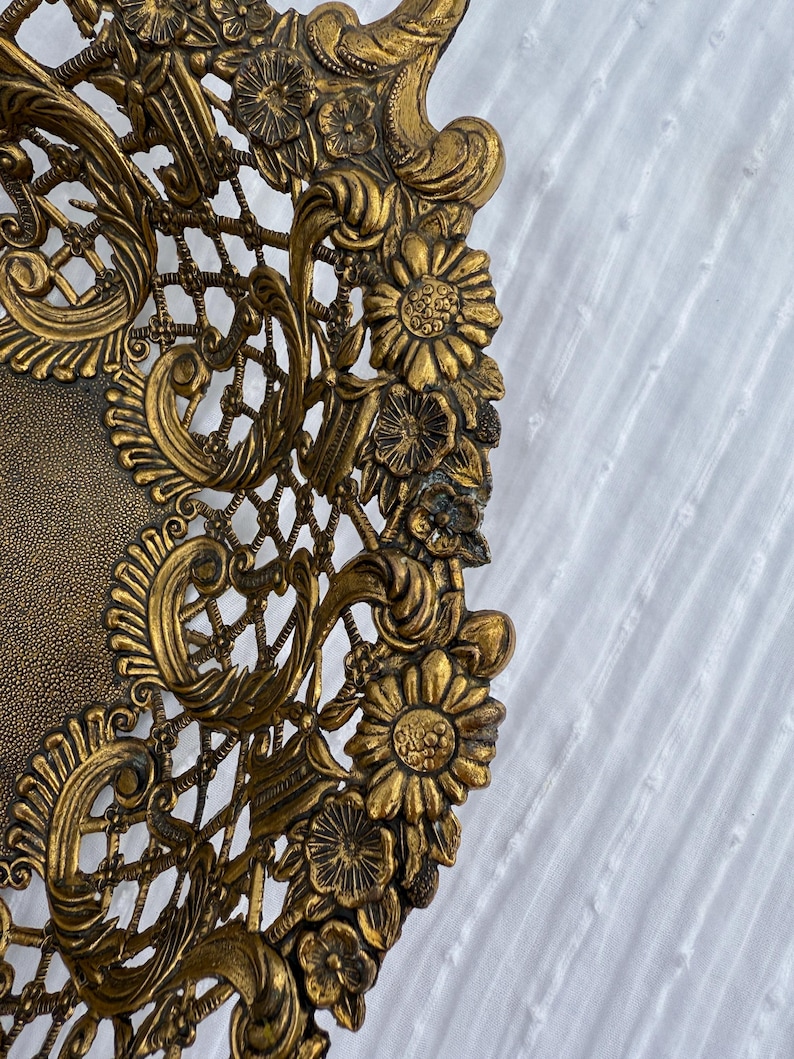 May include: Ornate gold-colored decorative plate with intricate floral and lattice detailing. The plate features a textured center and a detailed, raised border with flowers and swirling patterns. The design is symmetrical and the plate has an antique appearance.