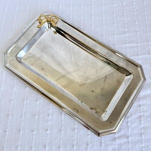 Puede incluir: Una bandeja rectangular plateada con un lazo dorado en una esquina. La bandeja tiene un borde elevado y una superficie pulida, adecuada para servir o exhibir.