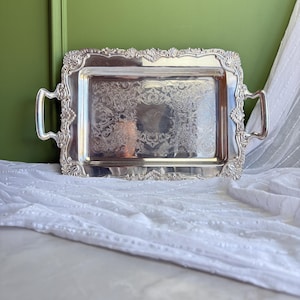 Puede incluir: Una bandeja de servicio rectangular de tono plateado con detalles en relieve en forma de concha a lo largo de los bordes y las asas. La superficie central de la bandeja muestra un intrincado patrón floral grabado con un acabado pulido y reflectante.
