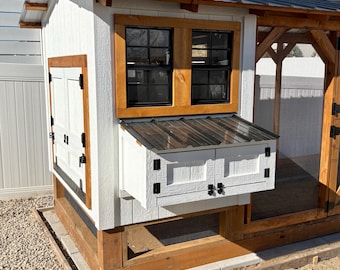 May include: A white and wood chicken coop with a metal roof. The coop has two black framed windows and a nesting box with two doors. The coop is built on a wooden frame and sits on a gravel base.