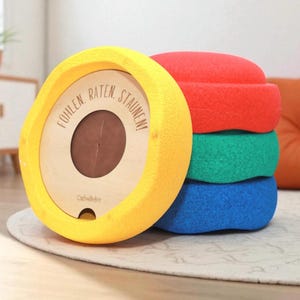 Può includere: Un giocattolo circolare giallo con la scritta "FÜHLEN. RATEN. STAUNEN!" su una faccia di legno. Dietro sono impilate tre forme arrotondate, strutturate e colorate in rosso, verde e blu. Il giocattolo è su un tappeto chiaro.