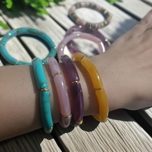 Op de afbeelding: Een verzameling kleurrijke armbanden in tinten turquoise, roze, paars en geel. De armbanden hebben gouden accenten en worden op een arm getoond. De armbanden zijn gemaakt van verschillende materialen en hebben een gladde, gepolijste afwerking.