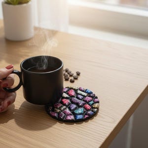 May include: A black mug filled with steaming coffee, resting on a colorful mosaic coaster. Coffee beans are scattered nearby. The mug is held by a person. A small potted plant sits in the background on a wooden table.