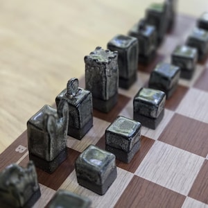 Puede incluir: Un juego de ajedrez con piezas de cerámica gris oscuro dispuestas sobre un tablero de ajedrez de madera. Las piezas tienen un acabado texturizado y esmaltado, y el tablero presenta un patrón de cuadros marrones y beige. Las piezas tienen varias formas, incluyendo torres, peones y caballos.