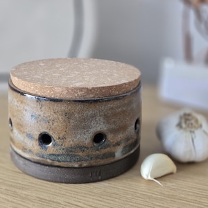 Op de afbeelding: Een keramische knoflookhouder met een kurken deksel. De houder is cilindrisch met een bruin en tan glazuur en verschillende ventilatiegaten. Een hele knoflookbol en een teentje liggen naast de houder.