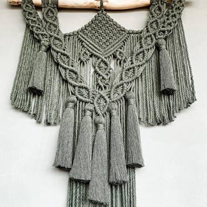 Könnte beinhalten: Handgefertigter Makramee-Wandbehang in Grautönen, aufgehängt an einem natürlichen Holzdübel. Das aufwendige Design zeichnet sich durch geknotete Muster, Quasten und fließende Fransen aus und schafft ein strukturiertes Dekor im Bohème-Stil.