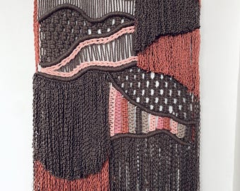 May include: A woven wall hanging with a wooden dowel at the top. The textile art piece features a layered design in shades of brown, pink, and gray, with fringe at the bottom. The piece has a textured, abstract pattern.