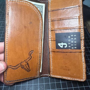 May include: Open, handmade leather wallet in shades of brown. The interior features card slots and a bill compartment holding a dollar bill. A steer skull design is etched on the front. The text "CUSTOM LEATHER SIGNS" is visible on a card.