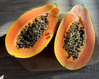 May include: Two halves of a ripe papaya fruit, showcasing vibrant orange flesh and a core filled with dark, round seeds. The fruit's skin is yellow, and the cut halves rest on a wooden cutting board, with natural light casting shadows.