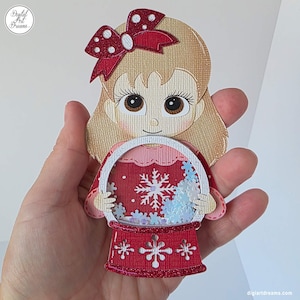 May include: A papercraft snow globe featuring a cartoon girl with blonde hair and a red bow. The snow globe has a red base with snowflake cutouts and a red interior with snowflakes and blue accents. The text "digiartdreams" is visible.