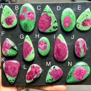 Peut inclure: Une collection de quatorze cabochons de zoïsite polis étiquetés de A à N, présentant chacun une base vert vif avec des inclusions de rubis magenta distinctes. Les pierres varient en forme, incluant des gouttes, des ovales et des triangles.