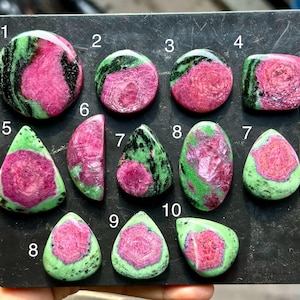 Könnte beinhalten: Eine Sammlung polierter Rubin-in-Zoisit-Edelsteine, angeordnet auf einer schwarzen Oberfläche, jeweils nummeriert von 1 bis 10. Die Steine zeigen leuchtend rosa und magentafarbene Zentren, umgeben von gesprenkelten grünen und schwarzen Einschlüssen.