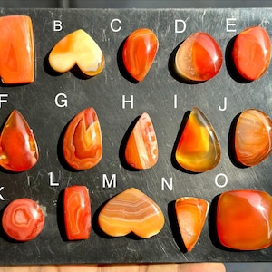 Può includere: Una collezione di quindici pietre preziose di corniola arancione e rossa lucidate, disposte su una tavola scura ed etichettate dalla A alla O. Le pietre presentano varie forme, tra cui rettangoli, cuori, gocce e cerchi con striature.