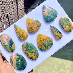 Puede incluir: Nueve piedras preciosas pulidas con patrones y colores únicos. Las piedras varían en forma, incluyendo ovaladas, en forma de lágrima e irregulares. Los colores incluyen tonos de verde, amarillo, naranja y rosa, con vetas y manchas intrincadas.