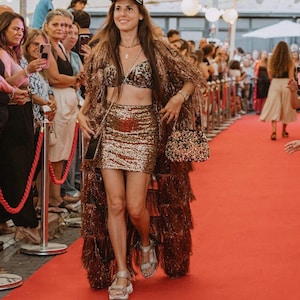 May include: A leopard print baseball cap, a patterned bralette, a gold sequined mini skirt, and a long bronze fringe duster coat. The outfit includes a small black and gold beaded handbag with a chain strap and metallic silver platform sandals.