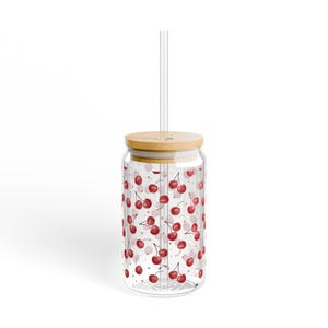 Könnte beinhalten: Ein durchsichtiger Glasbecher mit Bambusdeckel und einem durchsichtigen Strohhalm. Der Becher ist mit einem Muster aus roten Kirschen verziert. Der Becher ist für Getränke konzipiert.