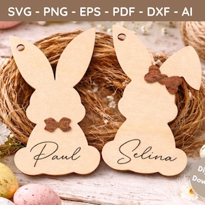Könnte beinhalten: Zwei hölzerne Hasen-Namensschilder mit braunen Schleifen, eines mit der Aufschrift "Paul" und das andere mit "Selina". Die Schilder sind vor dem Hintergrund eines geflochtenen Nestes, Ostereiern und Blumen platziert. Das Bild enthält den Text "SVG - PNG - EPS - PDF - DXF - AI".