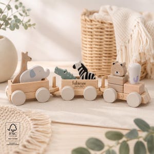 Könnte beinhalten: Ein hölzernes Spielzeugeisenbahn-Set mit drei verbundenen Waggons und hellen Rädern. Der Zug transportiert hölzerne Tierfiguren, darunter eine Giraffe, einen Elefanten, ein Krokodil, ein Zebra und einen Bären. Der mittlere Waggon ist mit dem Namen Fabienne und dem Datum 06.04.2025 graviert.
