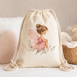 May include: A cream-colored drawstring backpack featuring a printed illustration of a fairy with pink wings and a pink dress, surrounded by green leaves and flowers. The name Mila is written in brown cursive script below the fairy figure.